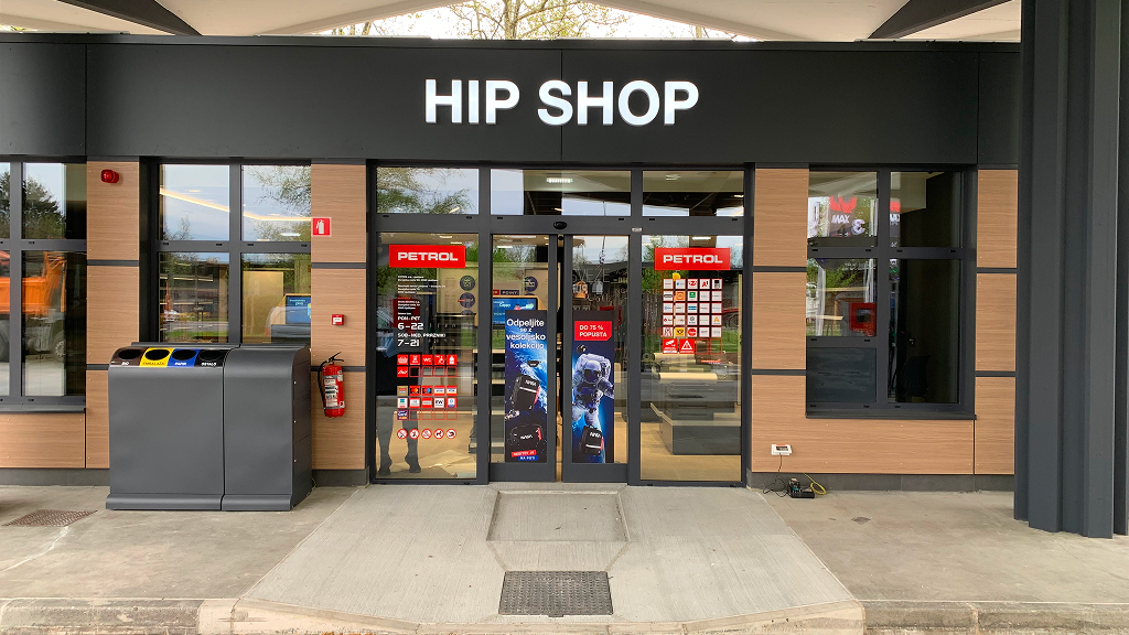 Vhod v prodajalno HIP SHOP na Petrolovem prodajnem mestu. Nad steklenimi vrati je velik osvetljen napis HIP SHOP, na vratih so Petrolove nalepke in promocijski plakati, ob vhodu pa stojijo zabojniki za ločeno zbiranje odpadkov.