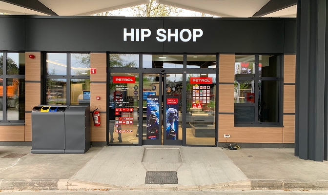 Vhod v prodajalno HIP SHOP na Petrolovem prodajnem mestu. Nad steklenimi vrati je velik osvetljen napis HIP SHOP, na vratih so Petrolove nalepke in promocijski plakati, ob vhodu pa stojijo zabojniki za ločeno zbiranje odpadkov.