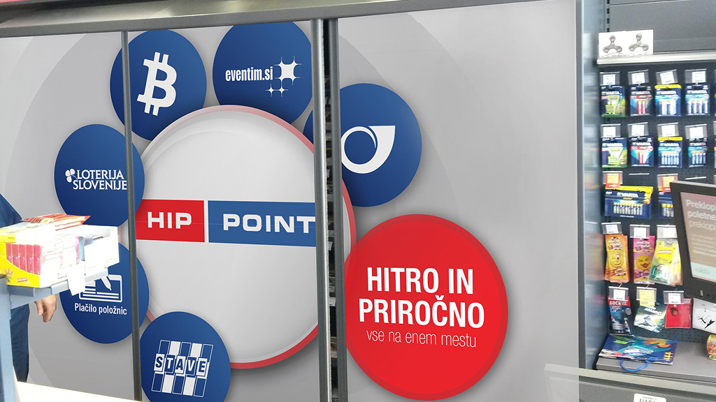 Nalepka HIP POINT na stekleni površini v prodajalni, z izpostavljenimi storitvami, kot so plačilo položnic, stave, Loterija Slovenije, eventim.si in druge hitre storitve. Na desni strani je rdeč krog z napisom »Hitro in priročno – vse na enem mestu«, ob njem pa je del prodajnega pulta z izdelki.