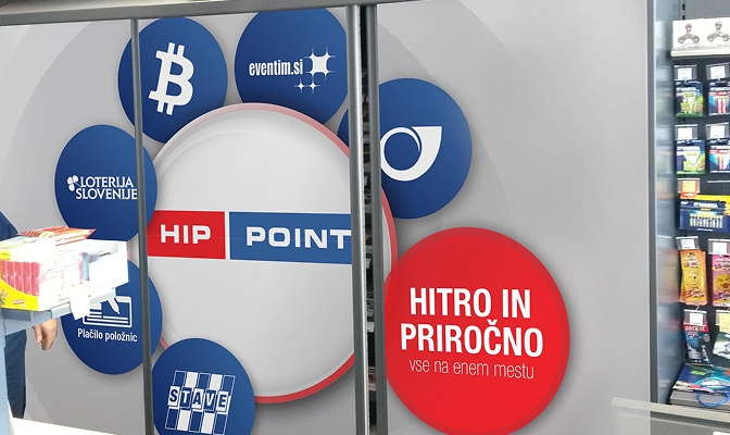 Nalepka HIP POINT na stekleni površini v prodajalni, z izpostavljenimi storitvami, kot so plačilo položnic, stave, Loterija Slovenije, eventim.si in druge hitre storitve. Na desni strani je rdeč krog z napisom »Hitro in priročno – vse na enem mestu«, ob njem pa je del prodajnega pulta z izdelki.