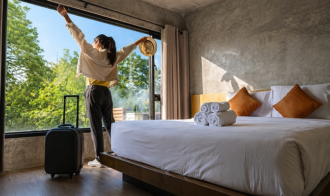 Na sliki je prikazana sodobno opremljena hotelska soba z veliko zakonsko posteljo, na kateri so zložene bele brisače in okrasne blazine v toplih zemeljskih tonih. Ob velikem panoramskem oknu stoji oseba z razširjenimi rokami, obrnjena proti naravi in jutranji svetlobi, v roki pa drži klobuk. Ob njej je postavljen kovček, kar nakazuje prihod ali odhod na potovanje. Skozi okno je viden zelen, sončen zunanji prostor z drevesi. Prizor ustvarja občutek sprostitve, udobja in brezskrbnega dopustniškega vzdušja.