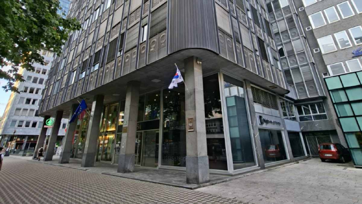 Na sliki je poslovna stavba s steklenim vhodom ter fasado iz kovine in stekla. Pred vhodom stojijo zastave na drogovih, ob stavbi pa so vidni tlakovana površina, drevesa in okoliške poslovne zgradbe.