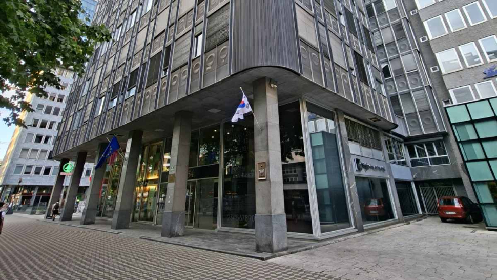 Na sliki je poslovna stavba s steklenim vhodom ter fasado iz kovine in stekla. Pred vhodom stojijo zastave na drogovih, ob stavbi pa so vidni tlakovana površina, drevesa in okoliške poslovne zgradbe.