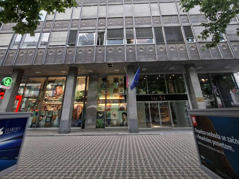 Na sliki je vhod v poslovno stavbo z velikimi steklenimi površinami in stebri. Nad vhodom je izobešena zastava Evropske unije. Na steklenih izložbah so vidni promocijski plakati, nad vhodom pa napis »biz 54«. Pred stavbo je tlakovana površina za pešce.