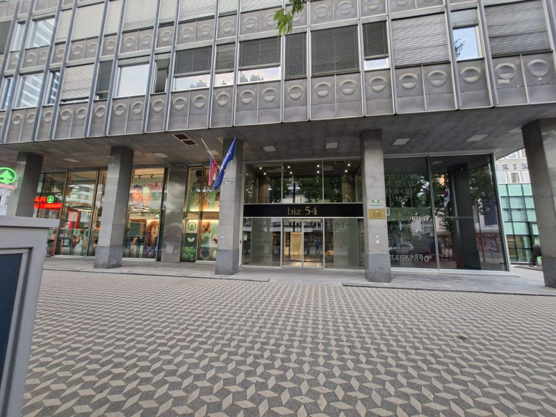 Na sliki je vhod v poslovno stavbo s steklenimi vrati in stebri. Nad vhodom je napis »biz 54«, ob vhodu pa visi zastava Evropske unije. Pred stavbo je tlakovana peš površina, ob straneh vhoda pa so velike steklene izložbe.