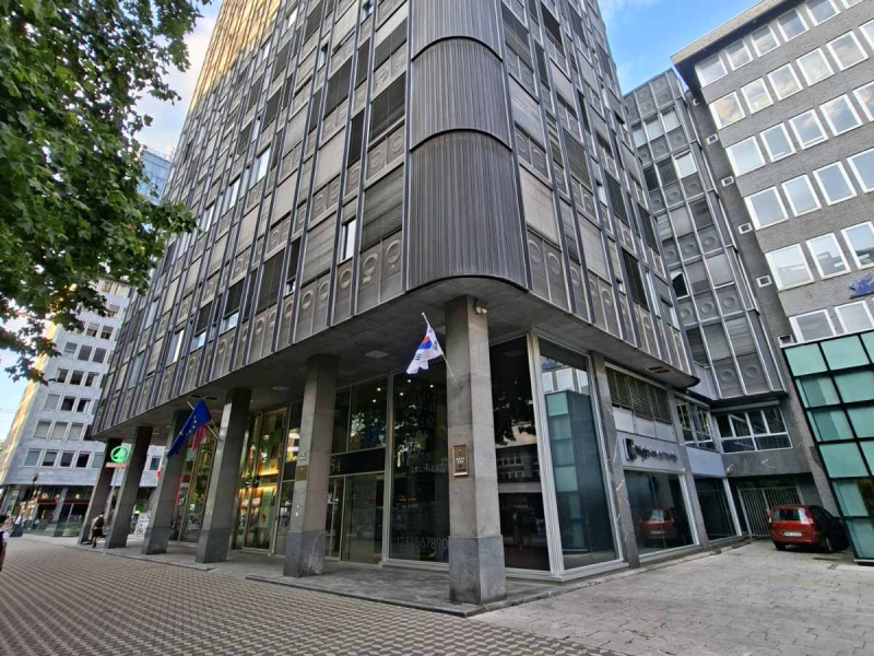 Na sliki je poslovna stavba s steklenim vhodom in visokimi stebri. Pred vhodom sta na drogovih izobešeni zastavi, ob stavbi pa so vidni tlakovana površina, okoliške poslovne zgradbe in parkirani avtomobili.