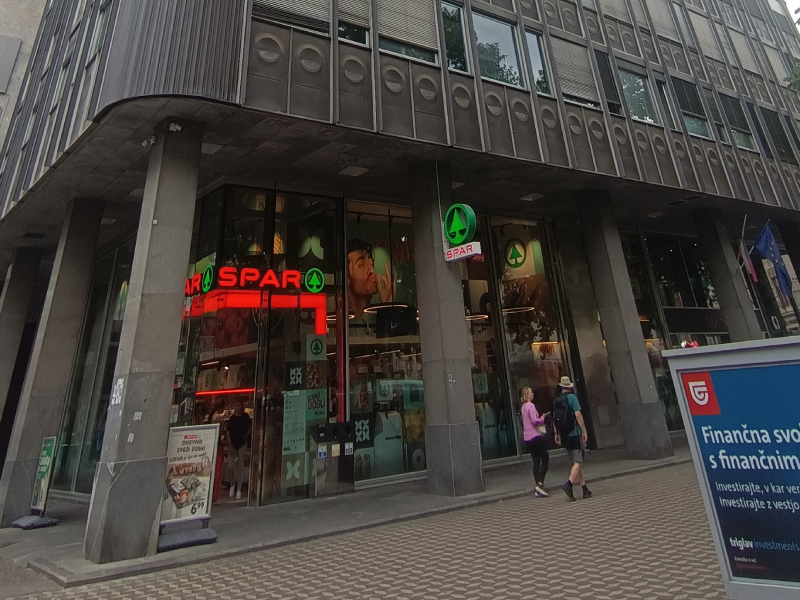 Na sliki je vhod v trgovino SPAR v pritličju poslovne stavbe. Nad vhodom so vidni osvetljeni logotipi SPAR, ob vhodu pa stojita dve osebi. Pred stavbo je tlakovana peš površina, na steklenih površinah trgovine pa so vidni promocijski plakati.