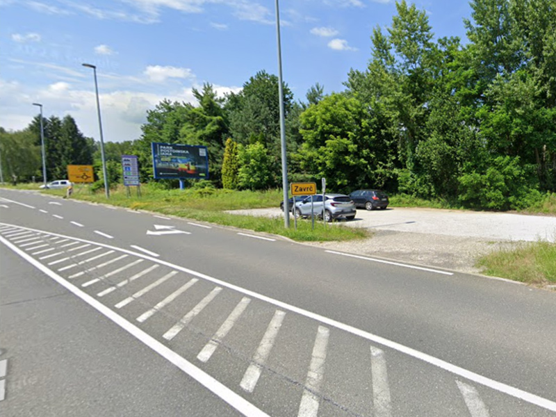 Na sliki je prikazano zemljišče ob prometni cesti z urejenim dovozom in makadamsko površino, obdano z zelenjem in drevesi.