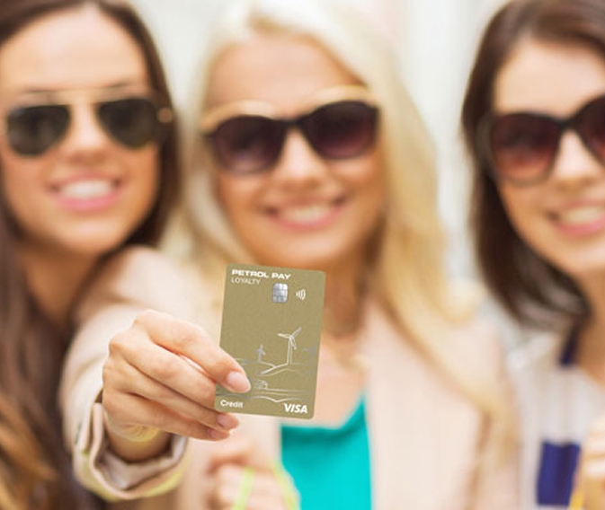 Tri nasmejana dekleta s sončnimi očali, ena v ospredju drži plačilno kartico Petrol Pay Loyalty Visa.