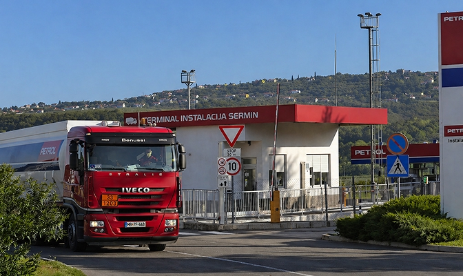 Na sliki je prikazan vhod v Petrolov terminal Terminal Instalacija Sermin. V ospredju po dovozni cesti vozi rdeč tovorni cisternski tovornjak Iveco, ki zapušča območje terminala. Ob vhodni stavbi so vidni prometni znaki in varnostna infrastruktura, v ozadju pa se razprostira zeleno gričevnato območje pod jasnim modrim nebom.