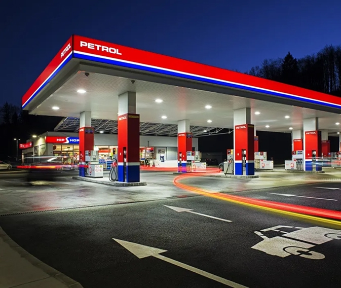 Na sliki je prikazan Petrolov bencinski servis v večernem času z osvetljenimi točilnimi mesti za gorivo in trgovino ob servisu.