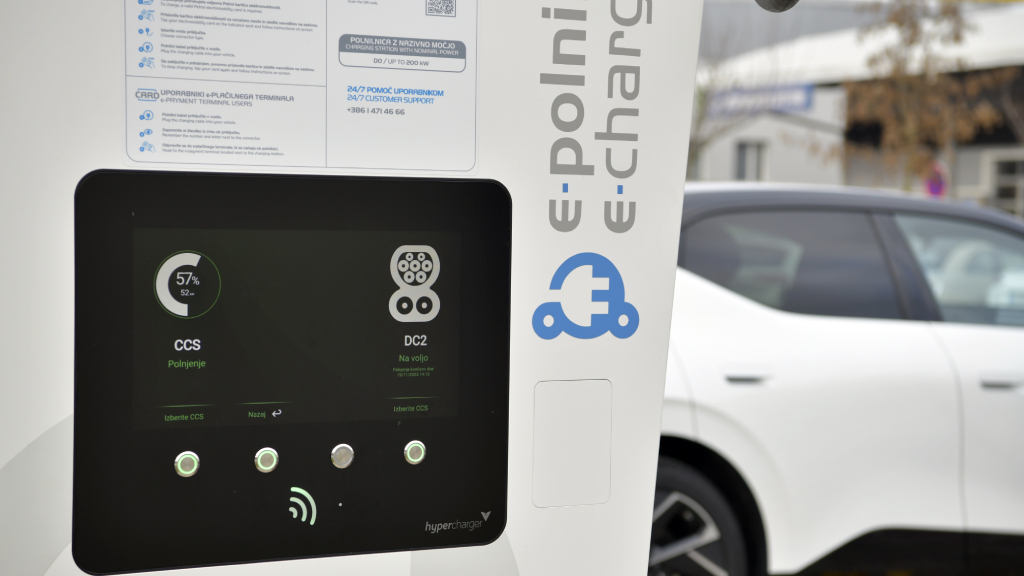 Na sliki je polnilnica za električna vozila z zaslonom za upravljanje postopka polnjenja, v ozadju pa je parkirano električno vozilo.