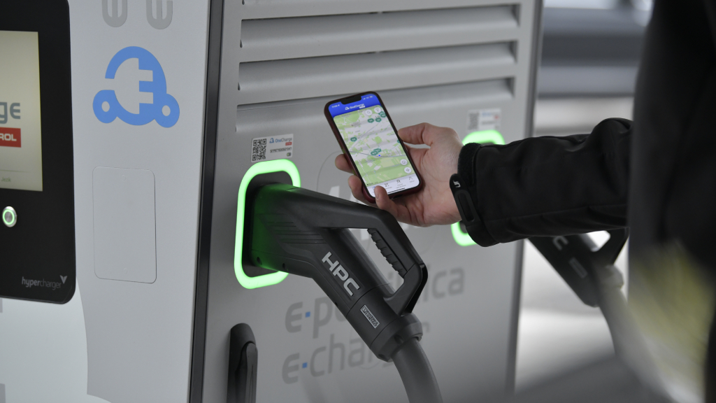 Na sliki je uporabnik, ki z mobilnim telefonom uporablja aplikacijo za iskanje in upravljanje polnjenja električnega vozila ob hitri polnilnici. V ospredju je prikazan polnilni priključek HPC, priključen na polnilno postajo za električna vozila.