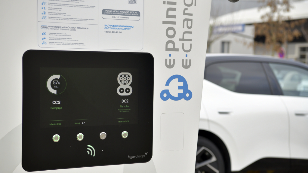 Na sliki je zaslon hitre polnilnice za električna vozila, ki prikazuje stanje polnjenja prek priključka CCS. Ob zaslonu je vidna polnilna postaja Petrol eCharge, v ozadju pa parkirano električno vozilo med postopkom polnjenja.