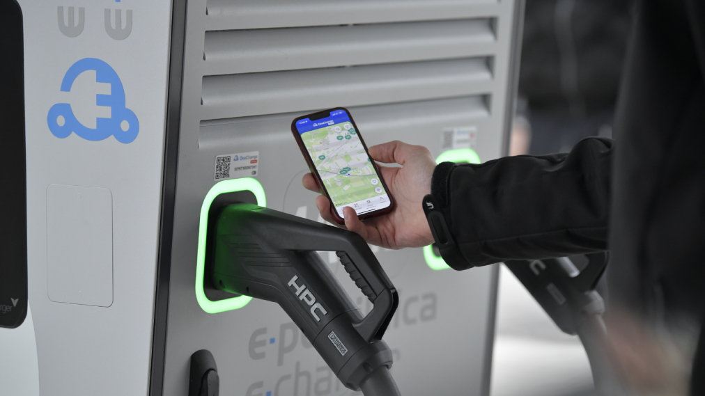 Na sliki je polnjenje električnega vozila na Petrolovi e-polnilnici, uporabnik pa ob polnilnici drži telefon z zemljevidom polnilnih mest.