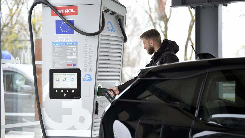 Moški polni električni avtomobil na Petrolovi e-polnilnici, nameščeni na bencinskem servisu, z vidnim polnilnim kablom in zaslonom polnilnice.