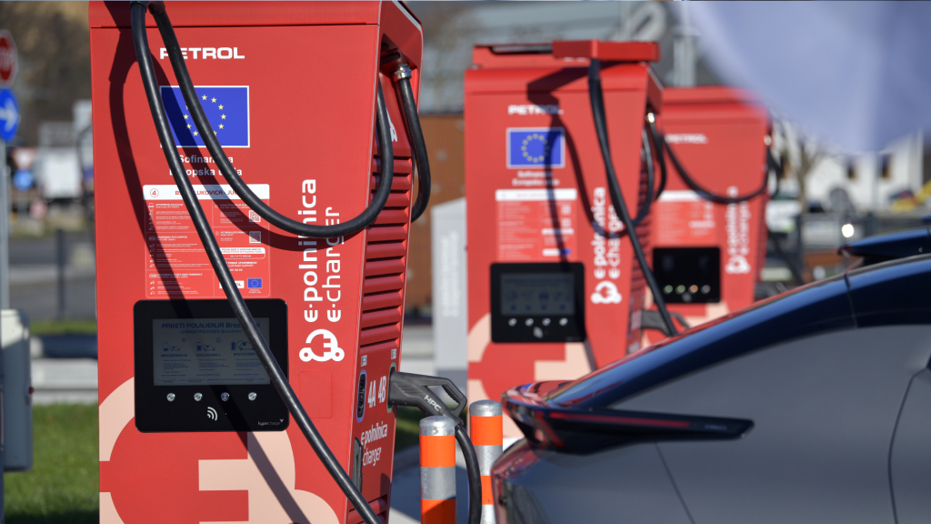Na sliki so hitre polnilnice Petrol eCharge za električna vozila, ob katerih je priključeno električno vozilo med postopkom polnjenja. V ospredju je prikazana polnilna postaja z zaslonom za upravljanje polnjenja in priključenim polnilnim kablom, v ozadju pa so vidne dodatne polnilnice v polnilnem parku.