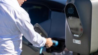 Oseba v srajci priklaplja moder polnilni kabel v električni avtomobil, v ospredju je sodobna e-polnilnica z vidnim zaslonom in oznakami.