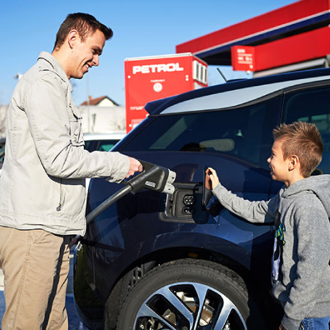 Moški in otrok stojita ob električnem avtomobilu ter priklapljata polnilni kabel na vozilo na Petrolovem prodajnem mestu, v ozadju je viden rdeč Petrolov objekt.