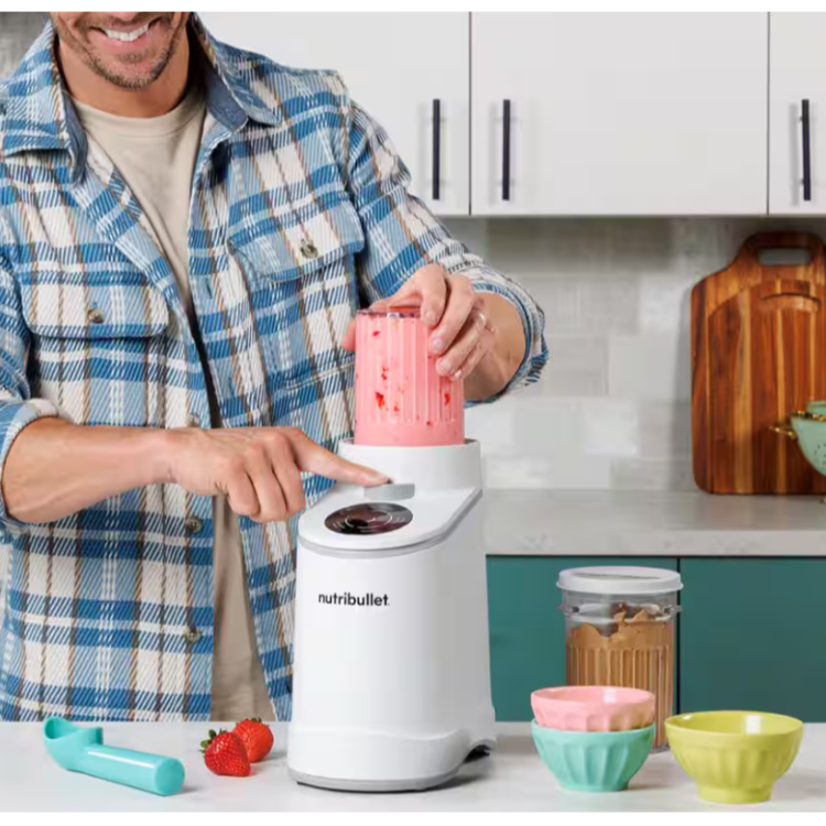 Oseba uporablja Nutribullet aparat za pripravo sladoleda/smoothijev, pritiska gumb na upravljalni plošči, posoda je napolnjena z rožnato zmesjo, na kuhinjskem pultu so sklede in sadje