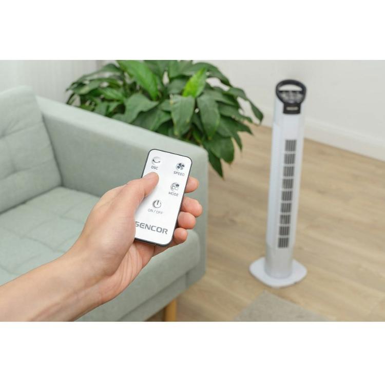 Sencor stolpni ventilator z daljinskim upravljalnikom v roki, postavljen v dnevnem prostoru