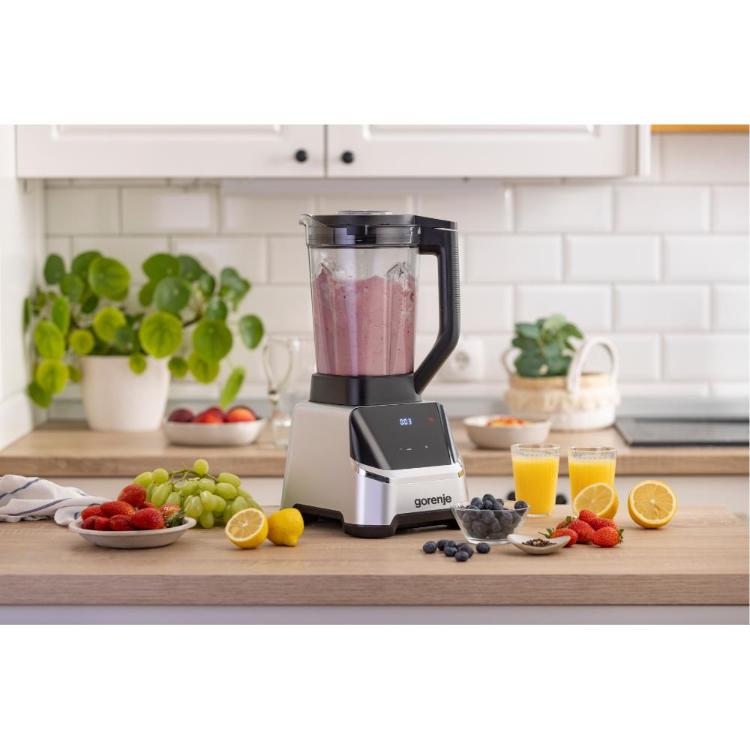 Na kuhinjskem pultu stoji moderni blender znamke gorenje. V njem je rožnata sadna zmes, na digitalnem zaslonu pa je prikazano »C03«. Okoli blenderja je razporejeno sveže sadje: jagode, zelene grozdne jagode, borovnice, limone in breskve. Na pultu sta tudi dva kozarca pomarančnega soka, majhna skodelica s poprovimi zrni in črtasta kuhinjska krpa. V ozadju so vidne posodice s sadjem in lončnice, ki ustvarjajo svetel, čist in svež kuhinjski ambient.