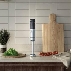 Electrolux palični mešalnik E5HB1‑6SS z inox mešalno nogo in ergonomskim ročajem, postavljen na kuhinjskem pultu ob zeliščih, paradižnikih in leseni deski za rezanje.