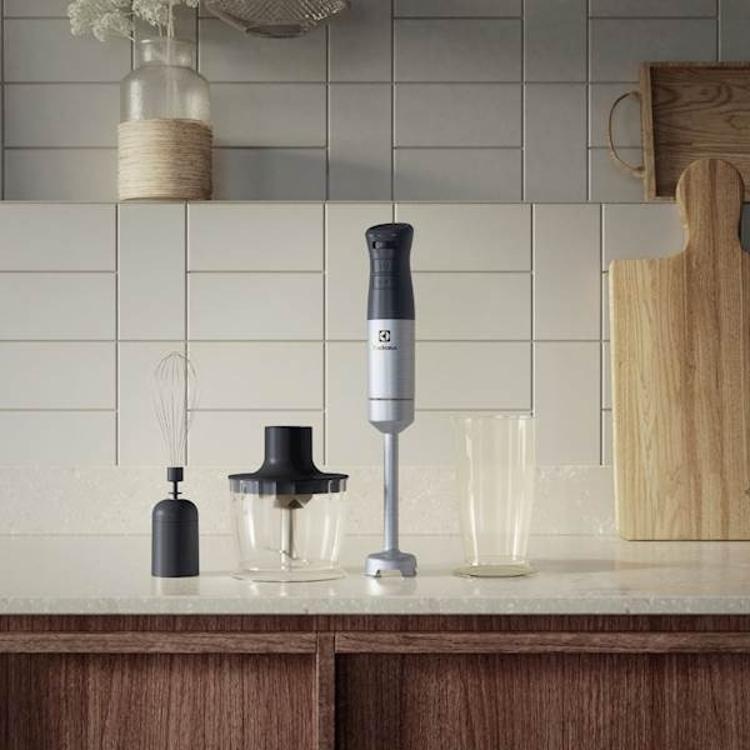 Electrolux palični mešalnik E5HB1‑6SS z nastavki za mešanje, stepanje in sekljanje, postavljen na kuhinjskem pultu ob merilni posodi in sekljalniku