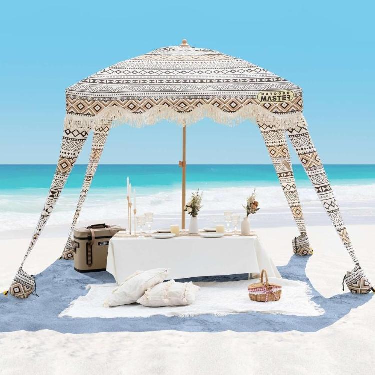Plažni paviljon z boho vzorcem in resastim robom, postavljen na peščeni plaži ob morju z ležalnim in piknik prostorom.