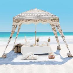 Plažni paviljon z boho vzorcem in resastim robom, postavljen na peščeni plaži ob morju z ležalnim in piknik prostorom.